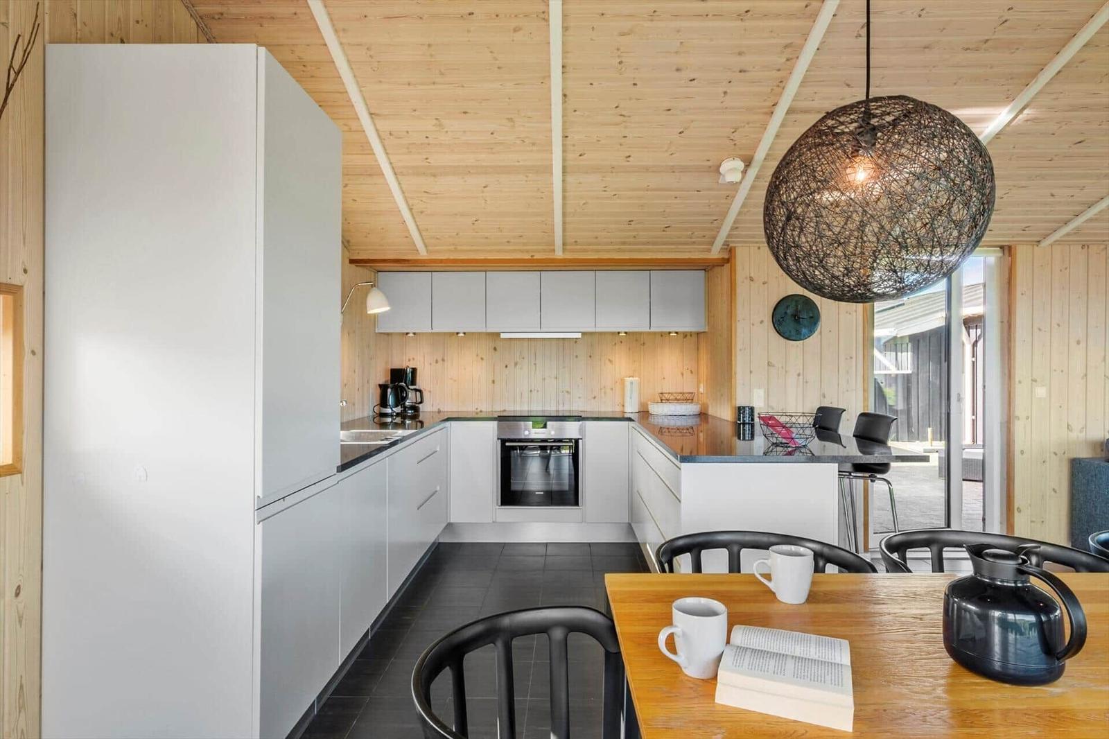 Küche mit Holzwand und -decke, weißer Einbauküche, Esstisch mit Stühlen und Hängelampe.