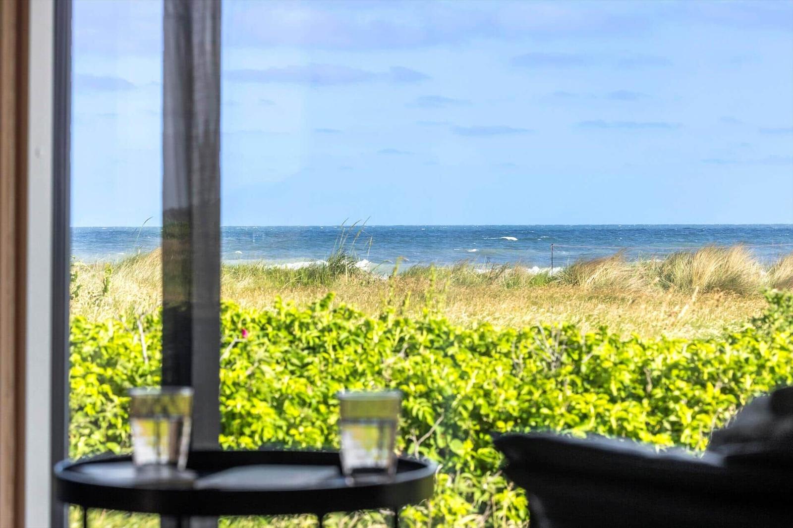 Durch das Fenster ist das Meer und Grünland zu sehen. Auf einem Tisch stehen zwei Gläser.