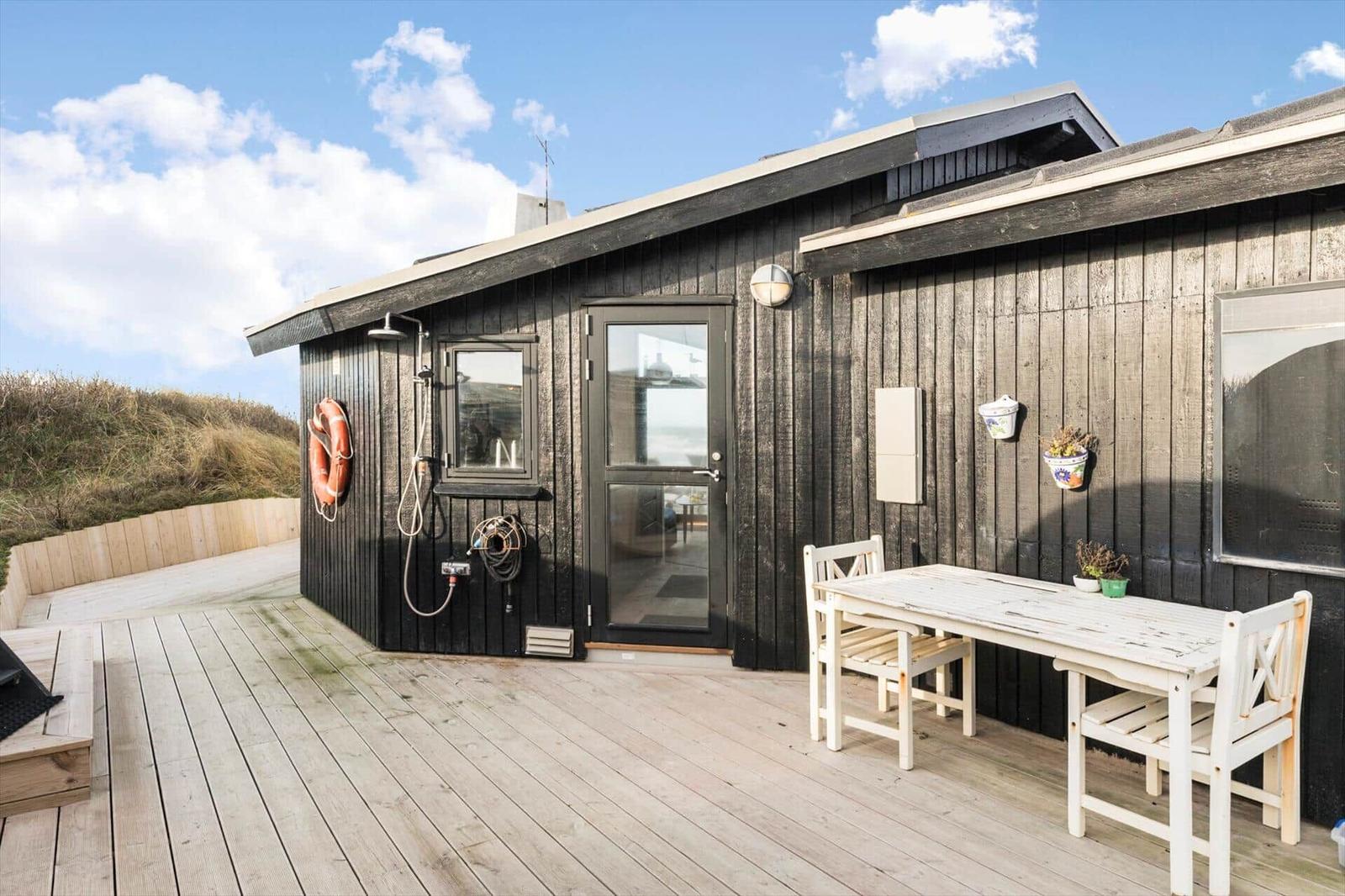 Holzterrasse mit Tisch, Stühlen und Außendusche an einem schwarz verputzten Haus.