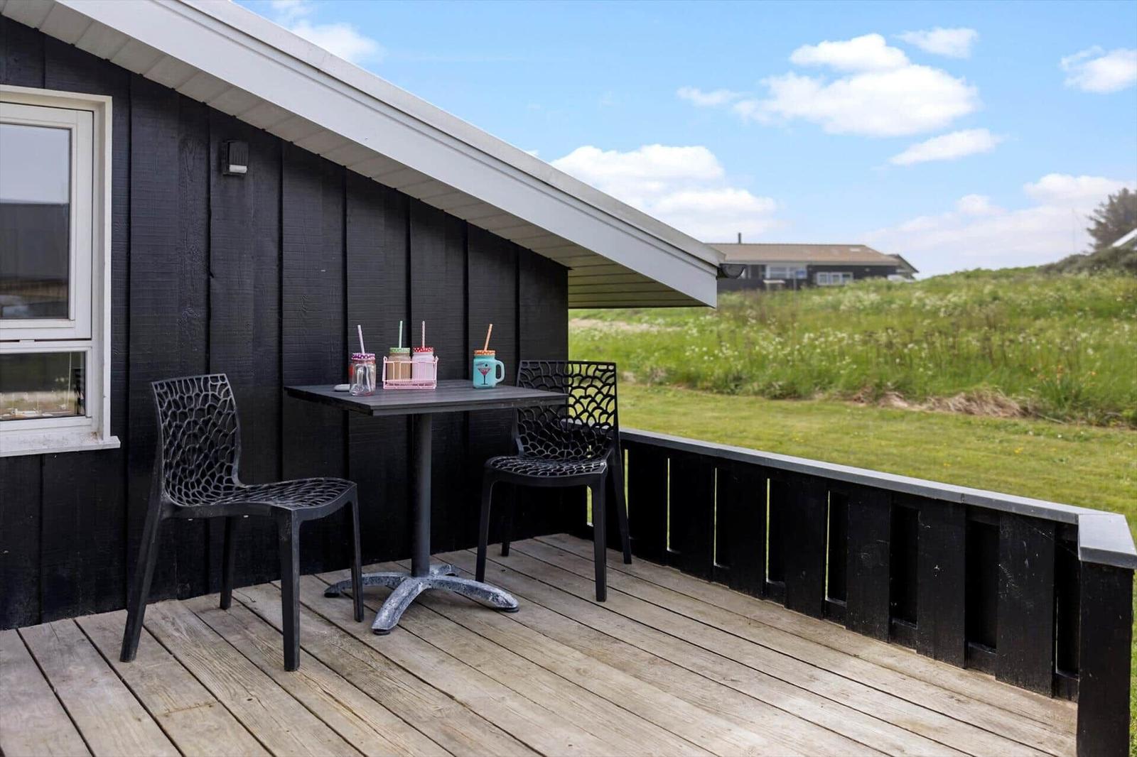 Terrasse mit Tisch und zwei Stühlen neben schwarzem Holzhaus mit Blick auf grünes Feld.