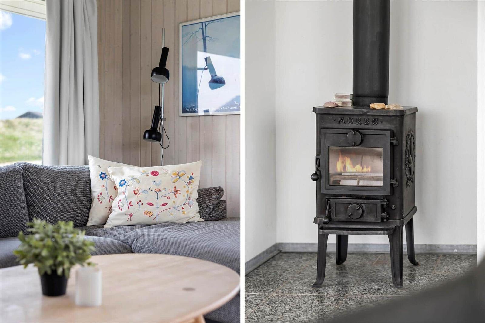 Grauer Sofa mit Stickereikissen, Holztisch und Wandbild. Kaminofen mit brennendem Feuer.