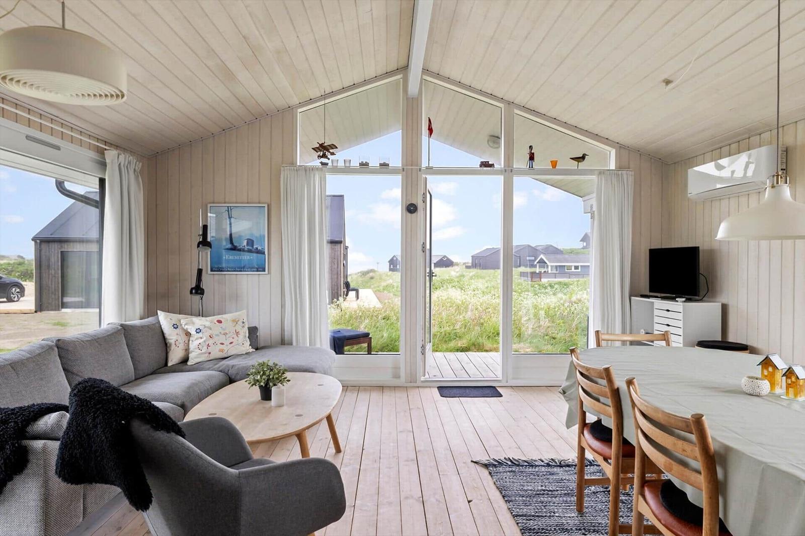 Wohn- und Essbereich mit großem Fenster, Sofa, Tisch und Stühlen. Draußen Grasland und Häuser.