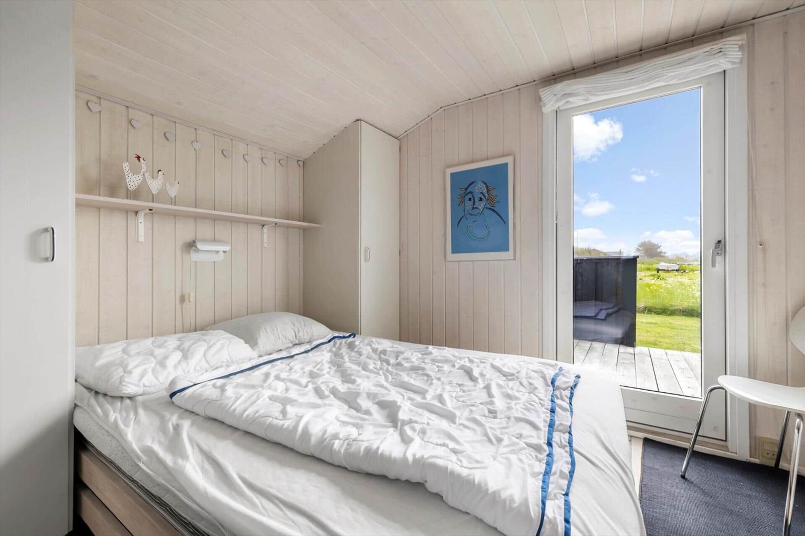 Schlafzimmer mit Bett, Fenster und Holzwand. Draußen ist eine Terrasse mit Blick auf Gras.