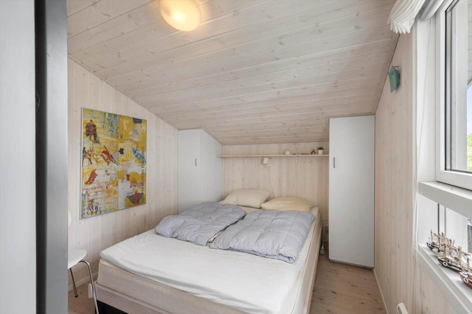 Schlafzimmer mit Bett, weißen Schränken und Holzwand. An der Wand hängt ein Bild.