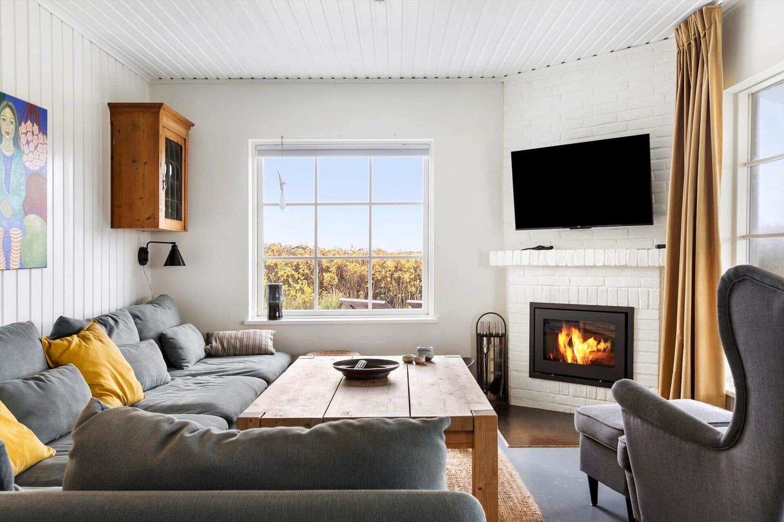 Wohnzimmer mit Sofa, Kamin und Fernseher. Fenster mit Blick auf Außengelände.