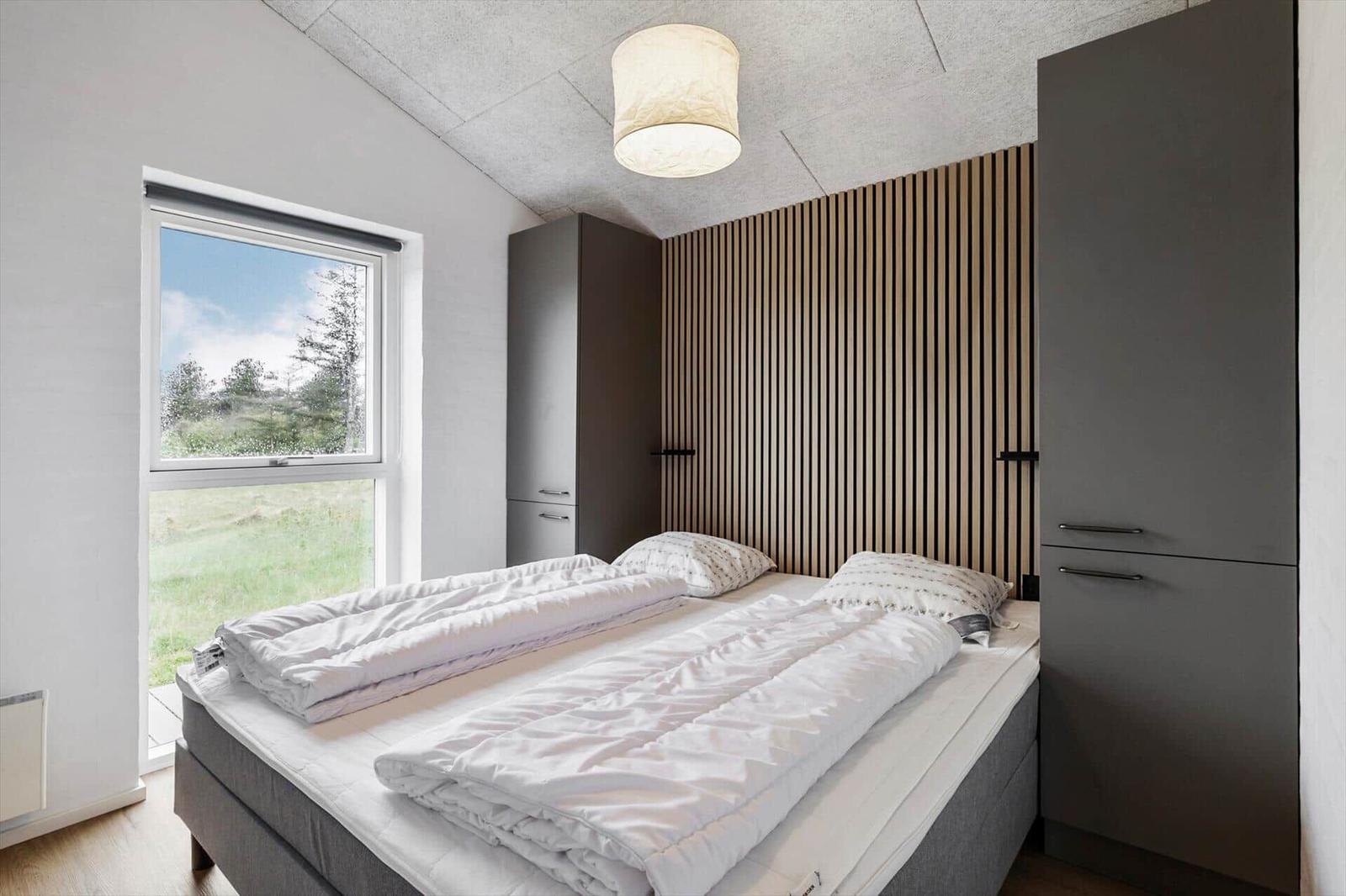 Schlafzimmer mit Doppelbett, Fenster und Holzwand. Zwei Schränke und Deckenleuchte.