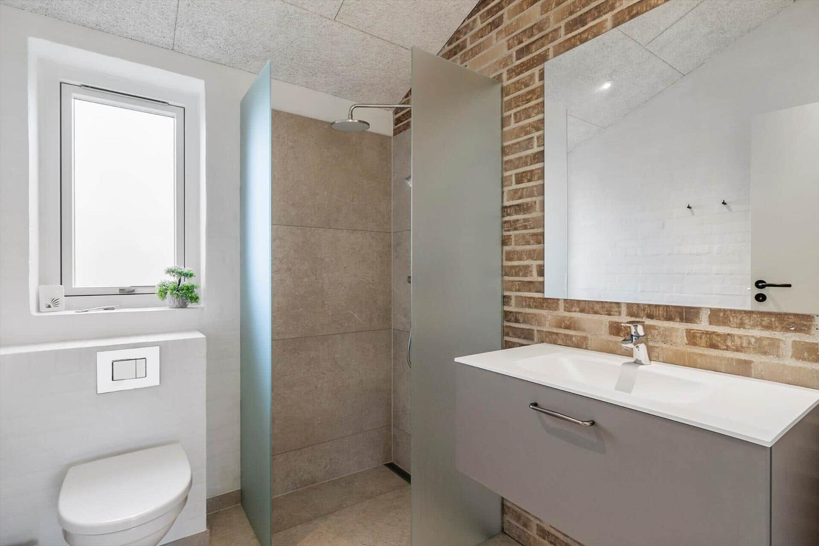 Bad mit Dusche, Waschbecken und Toilette. Wand aus Backstein und Fenster mit Pflanze.