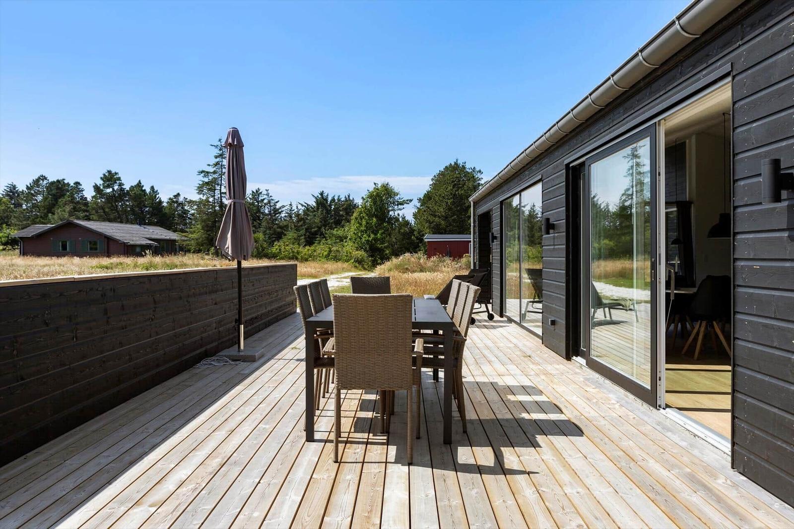 Terrasse mit Tisch, Stühlen und Sonnenschirm. Glasfront führt ins Haus. Blick auf Wald und Himmel.