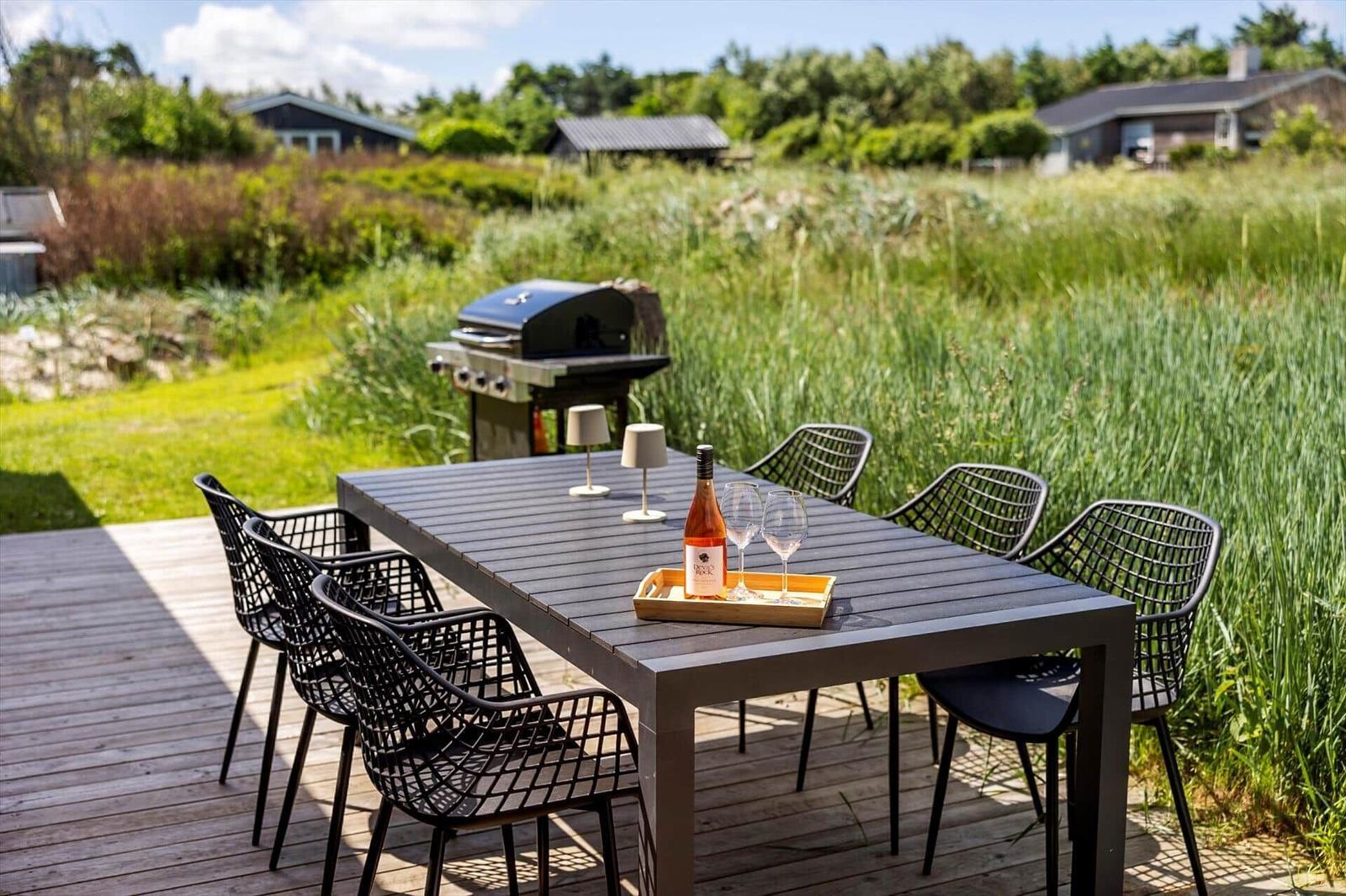 Holzterrasse mit Tisch, Stühlen, Grill und Flasche Wein.