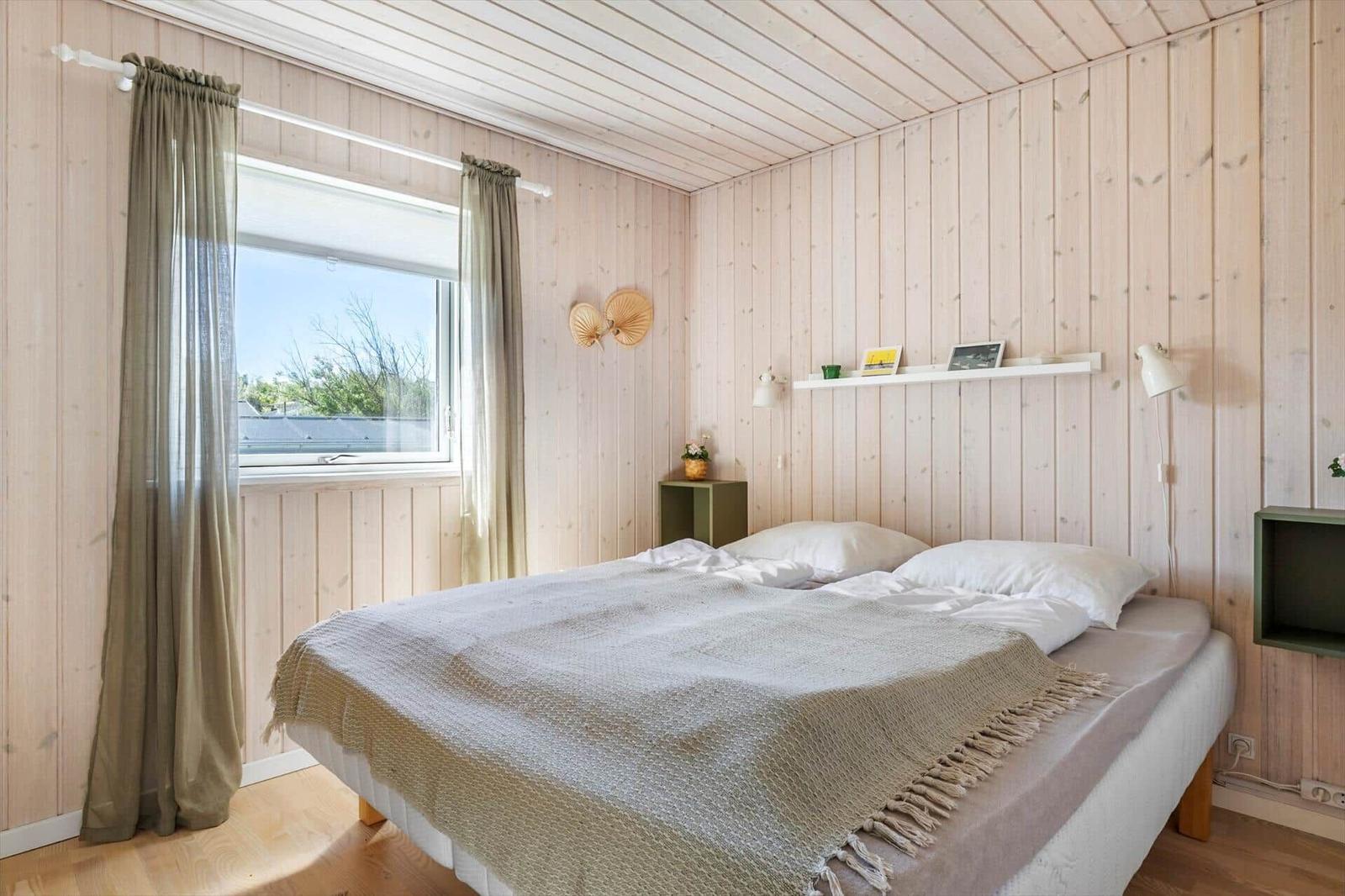 Schlafzimmer mit Holzwandverkleidung, Bett und Fenster mit Vorhängen.