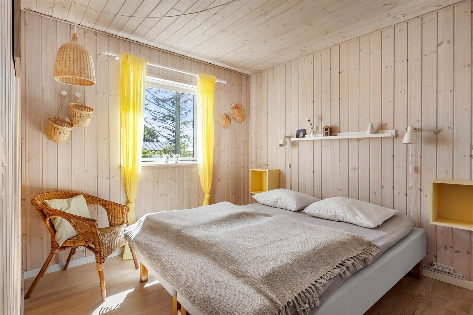 Schlafzimmer mit Holzwand, Bett, Fenster mit gelben Vorhängen und Stuhl.