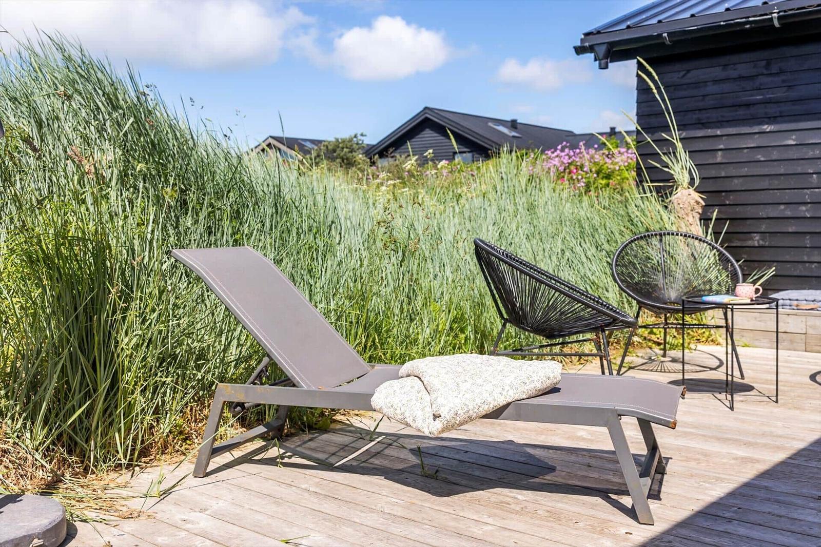 Terrasse mit Liegestuhl, zwei Stühlen und Tisch. Hintergrund: hohe Gräser und dunkle Holzwände.