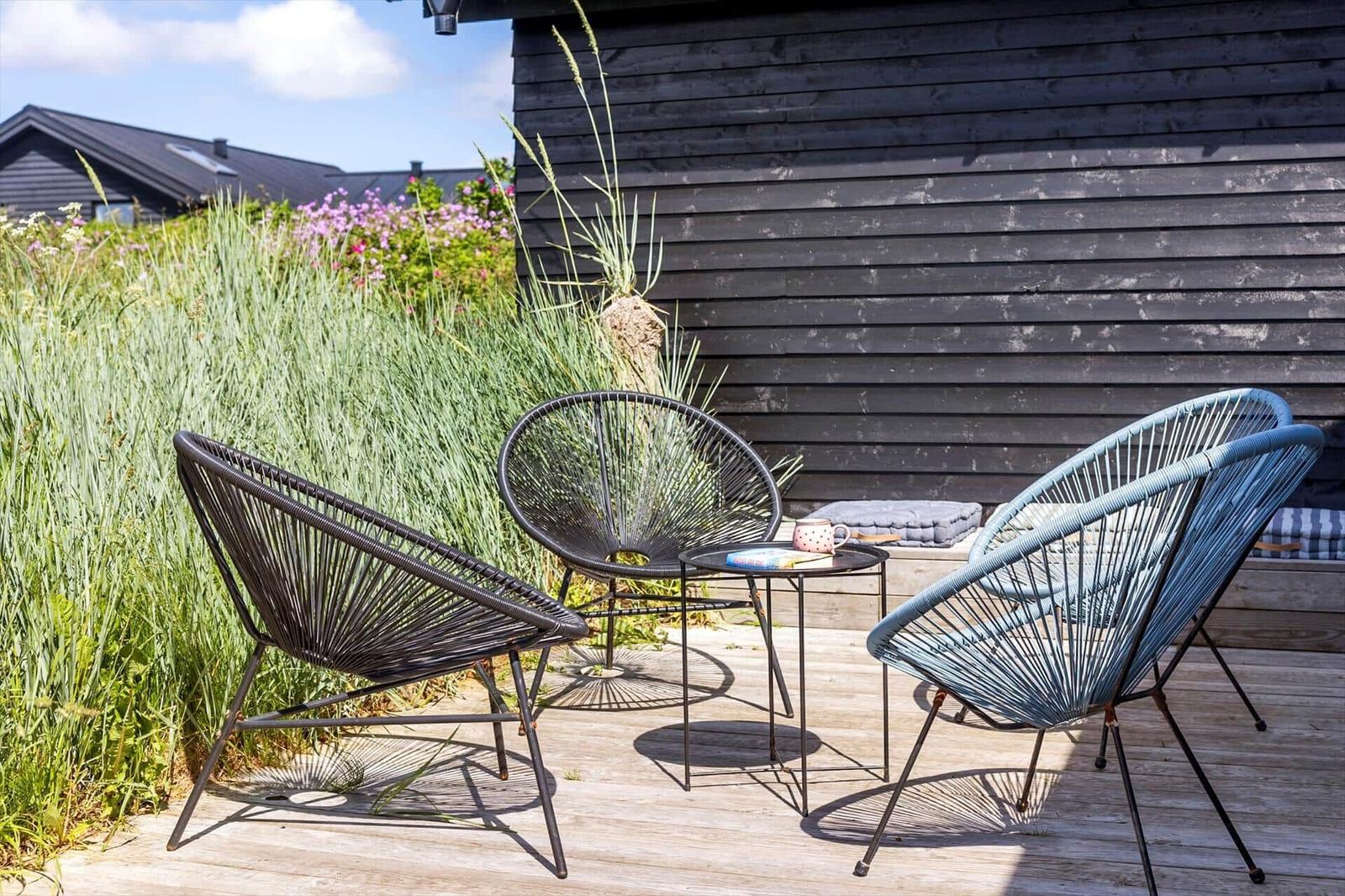 Drei Stühle und ein Tisch auf einer Terrasse vor einem Holzhaus mit Gras und Blumen.