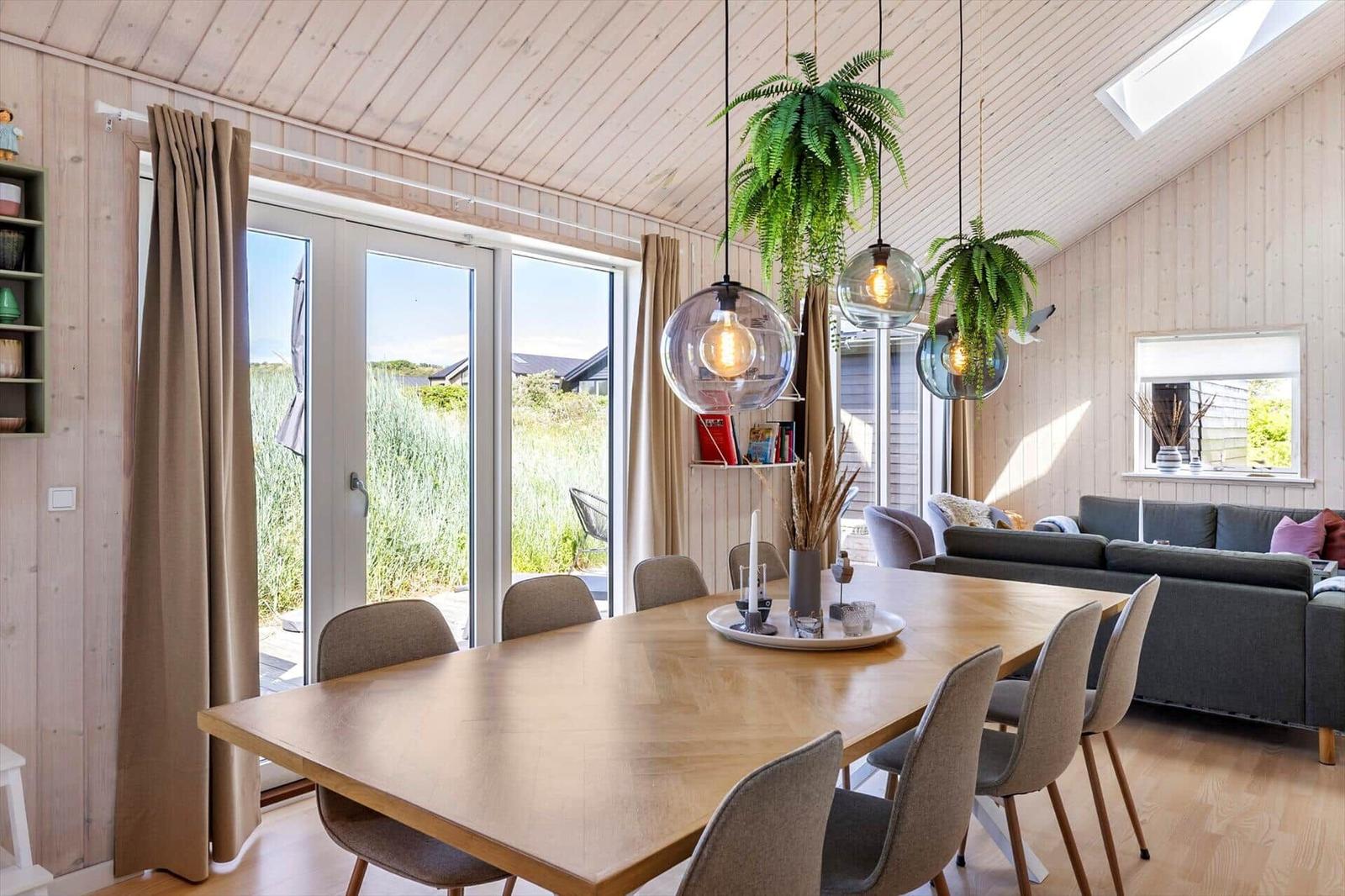 Essbereich mit Holztisch, Stühlen und Pflanzen. Durchgang zur Lounge mit Sofa und Fenster.