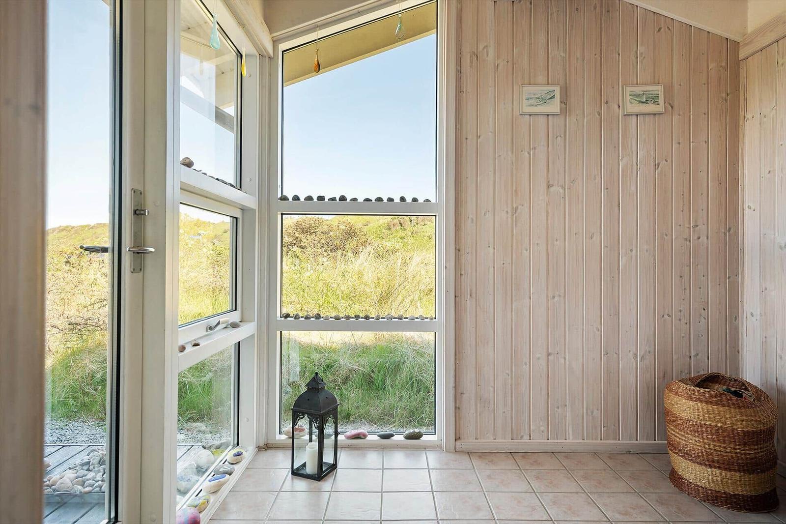 Raum mit Holzwand, großem Fenster und Blick auf Grasland. Bodenfliesen, Laterne und Korb.