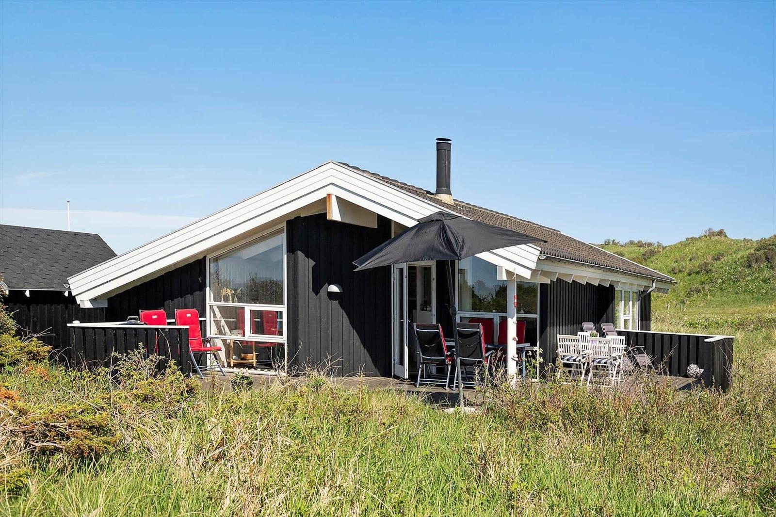 Schwarzes Holzhaus mit Terrasse, Sonnenschirm und Gartenmöbeln. Grün bewachsene Landschaft im Hintergrund.