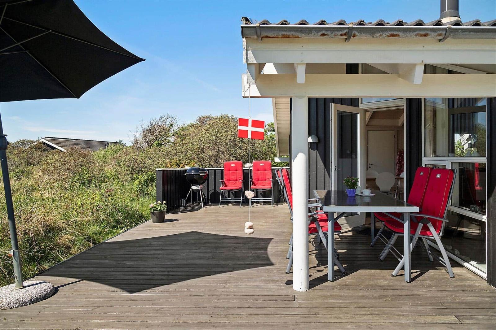 Terrasse mit Tisch, Stühlen und Grill unter schwarzem Sonnenschirm. Daneben Flagge und Pflanzen.