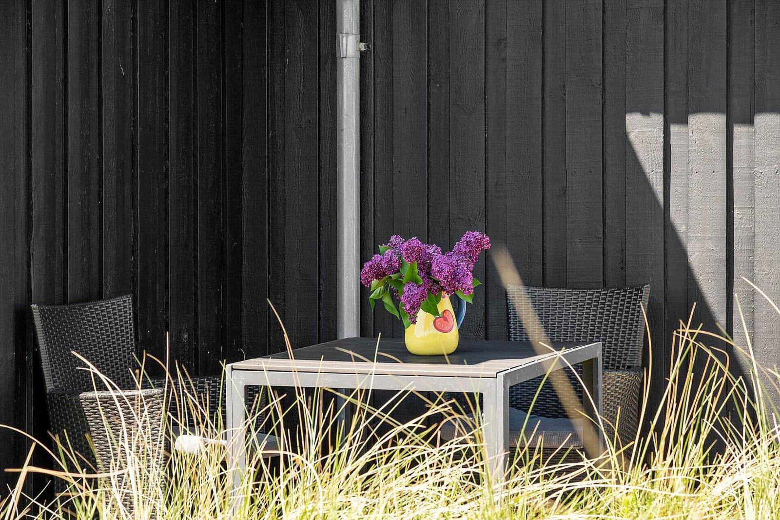 Außenbereich mit Tisch, zwei Stühlen und einer Vase mit lila Blumen vor schwarzem Holz.