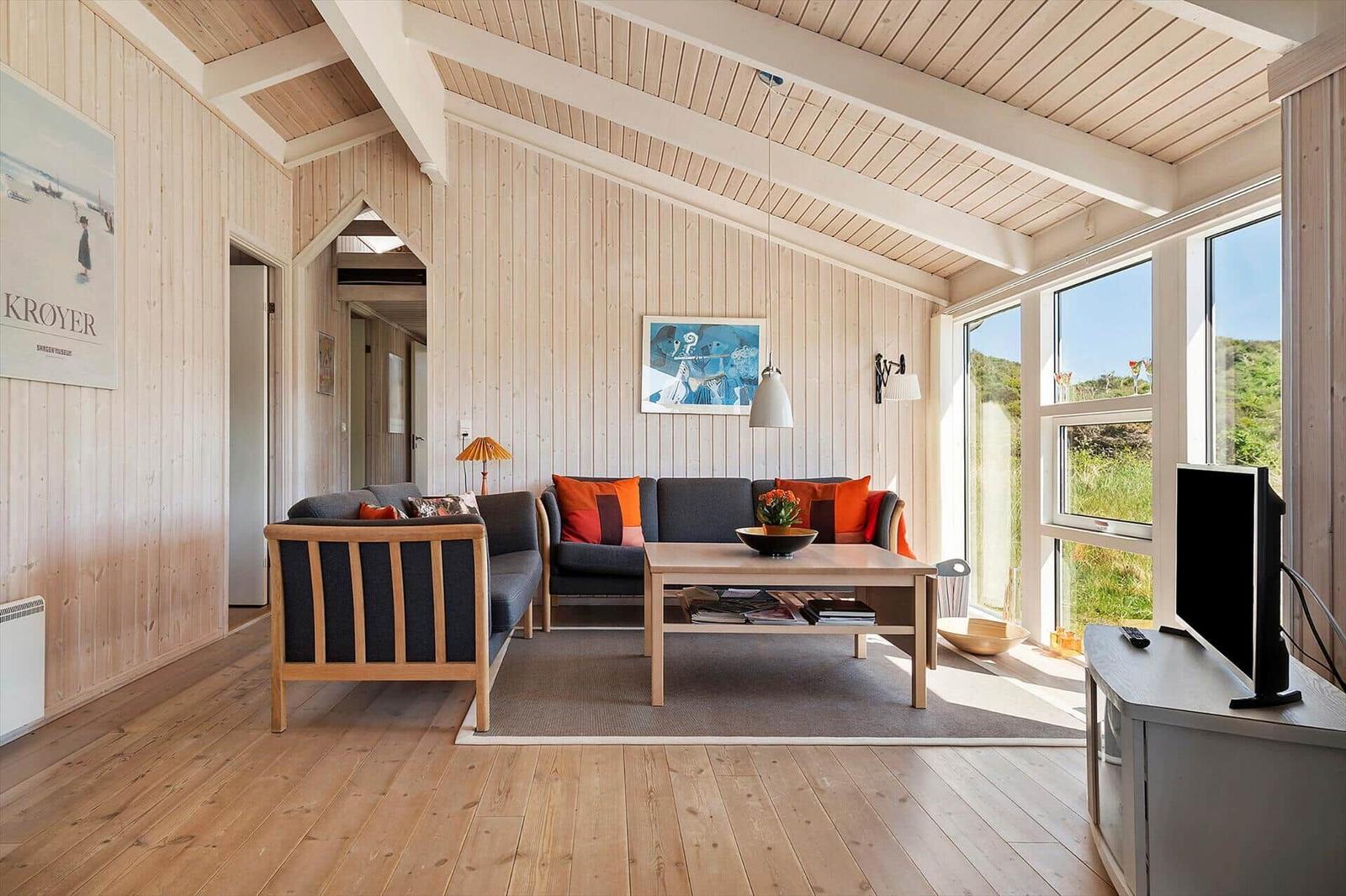 Wohnzimmer mit Holzwänden, Sofa, Couchtisch und TV. Große Fenster mit Blick auf Grünfläche.