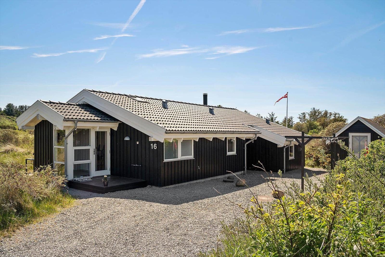 Ein schwarz-weißes Ferienhaus mit Nummer 16, umgeben von Gras und Sträuchern unter blauem Himmel.