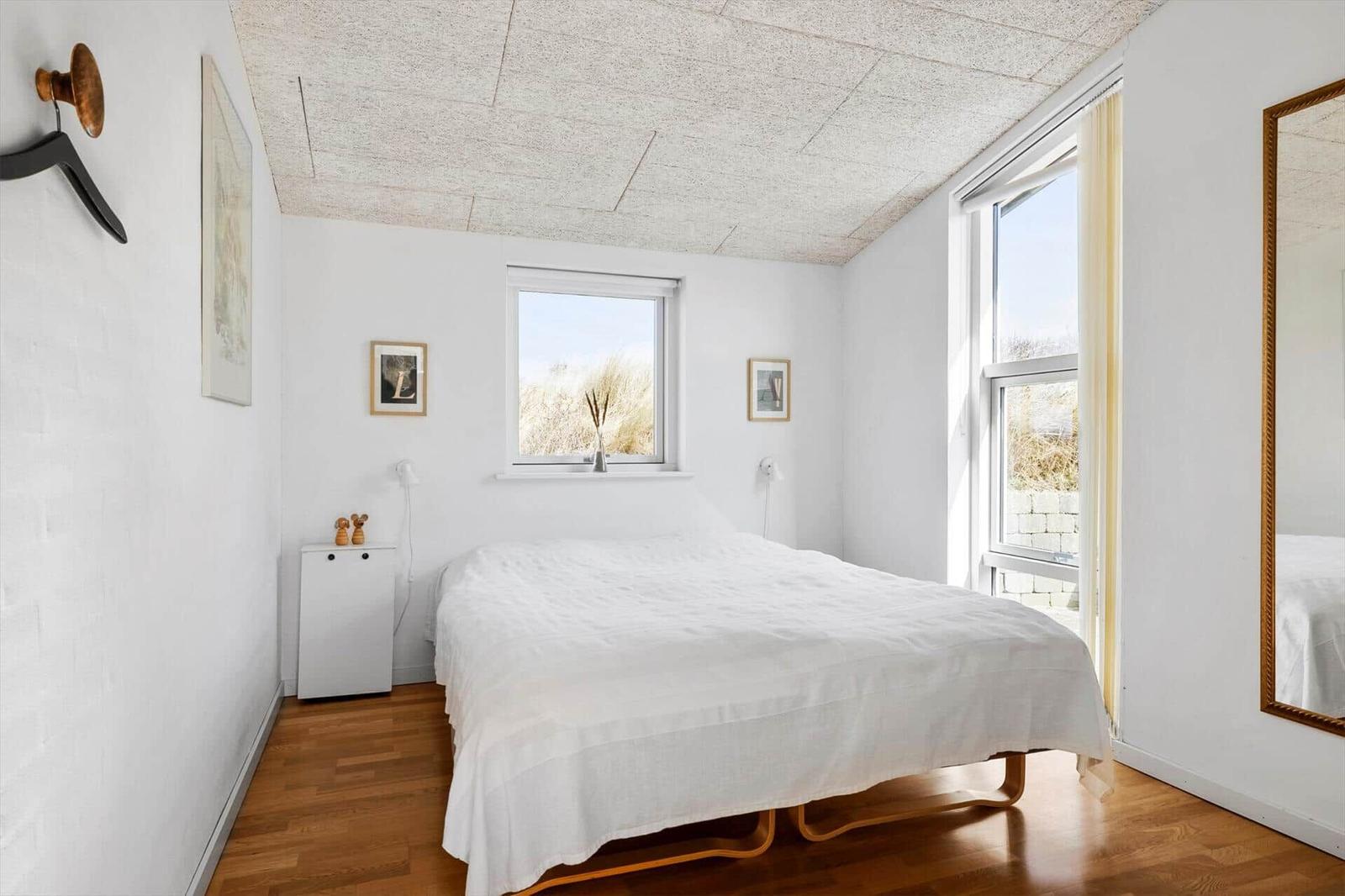 Schlafzimmer mit weißem Bett, Holzboden und großen Fenstern. Decke mit Paneelen.