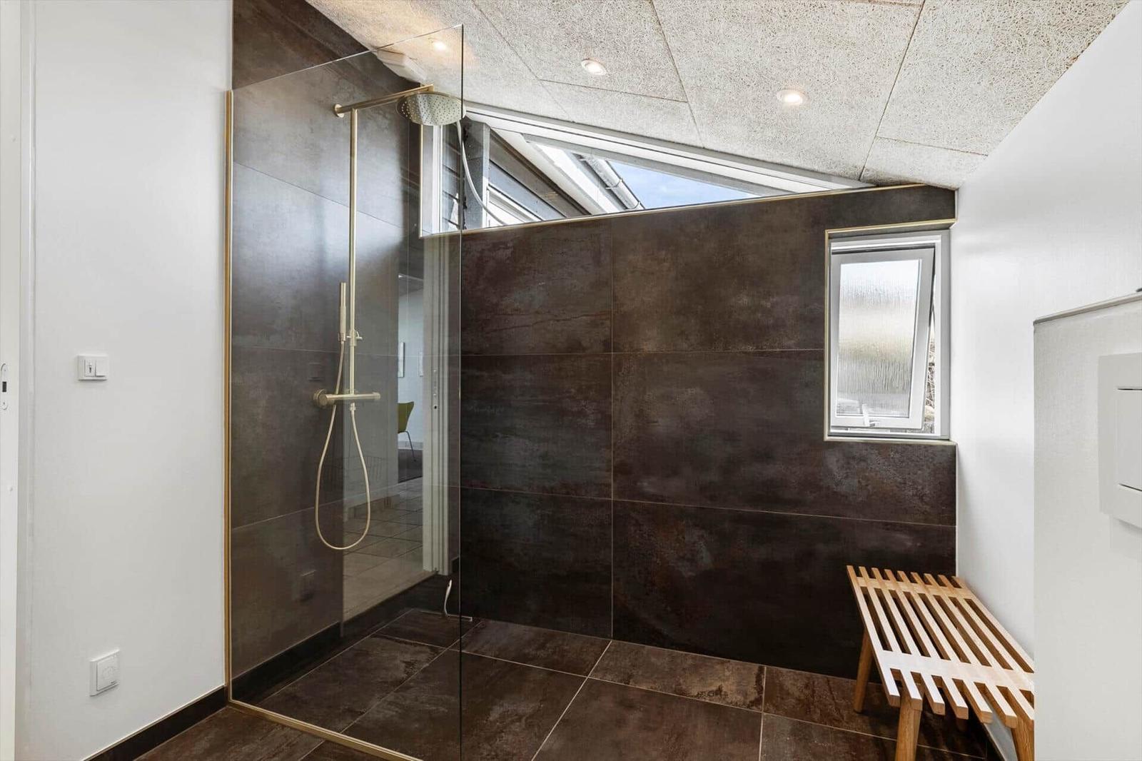 Dusche mit Glaswand, dunklen Fliesen und Holzbank. Fenster und Dachfenster sorgen für Licht.