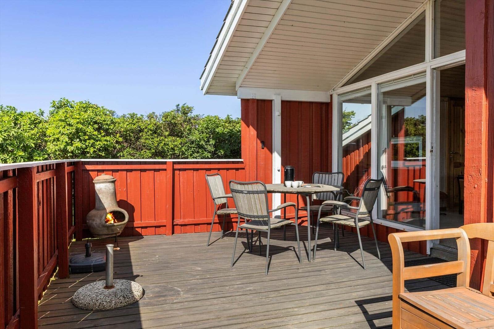 Terrasse mit Tisch, Stühlen und Feuerkamin. Roter Holzaufbau und grüne Vegetation im Hintergrund.