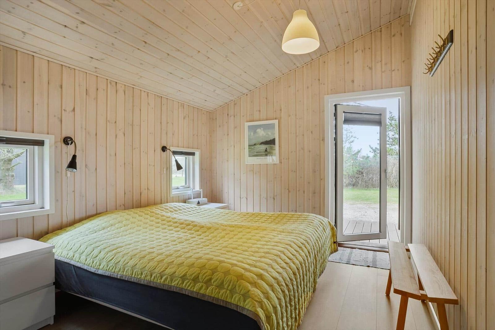 Schlafzimmer mit Holzwand, Bett mit gelbem Decken, Fenster und Tür zum Außenbereich.