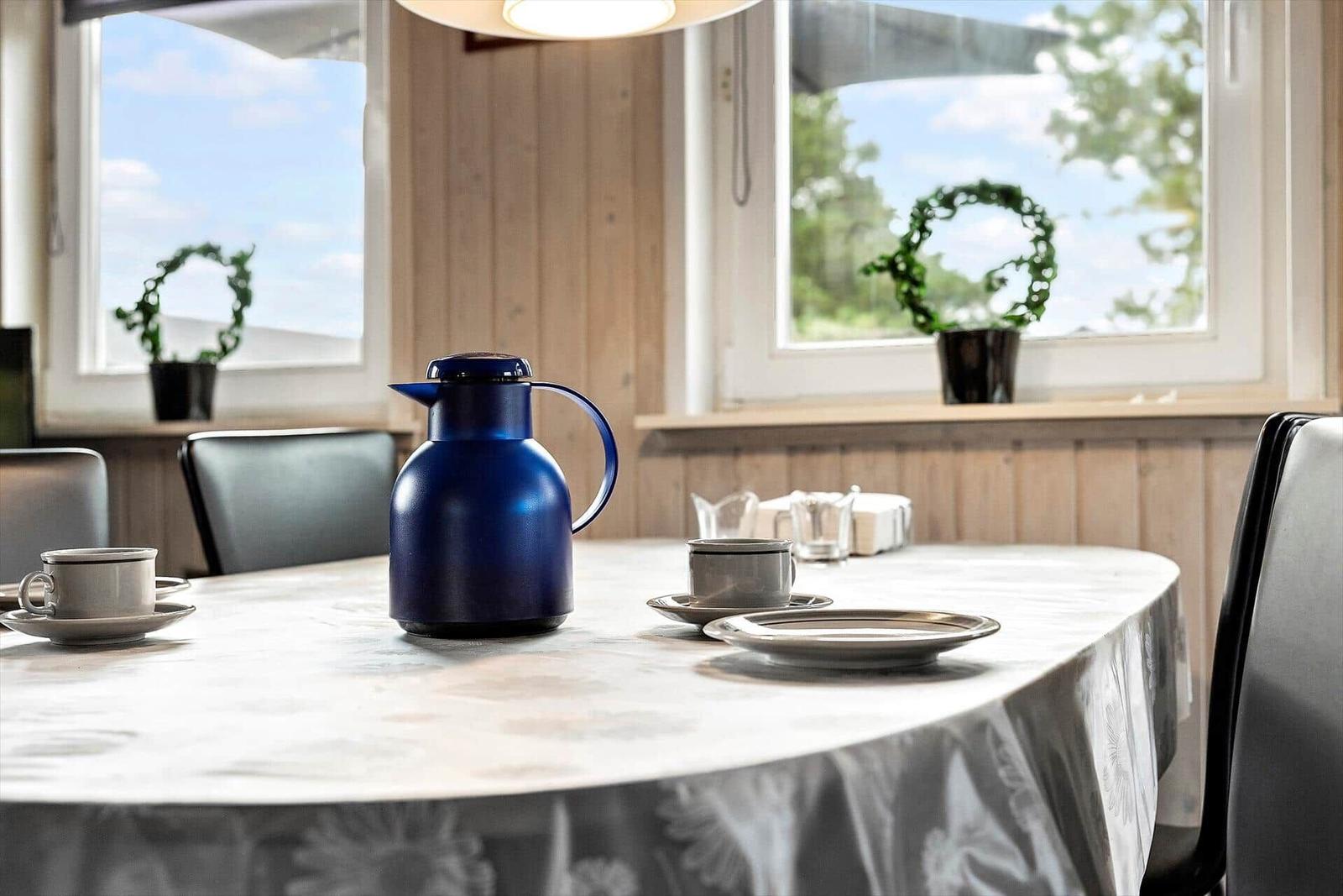Rundtisch mit Tischdecke, Thermoflasche und Tassen. Fenster mit Pflanzen im Hintergrund.