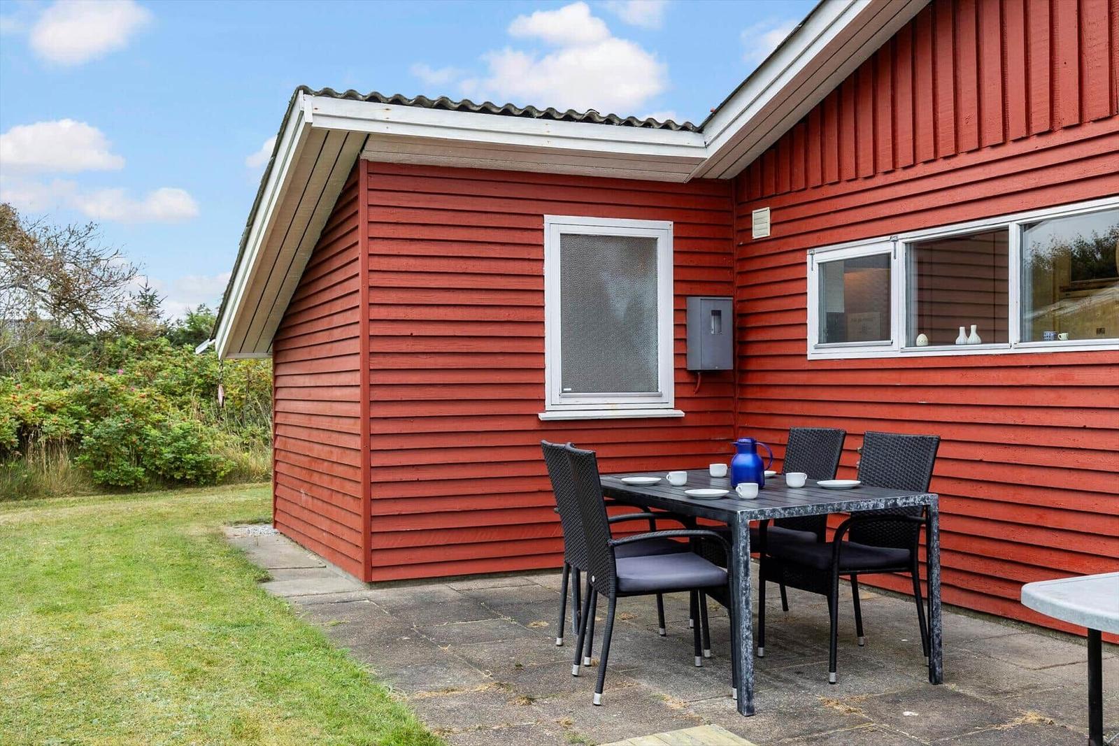 Rotes Holzhaus mit Terrasse, Tisch und Stühlen. Grasfläche und Bäume im Hintergrund.