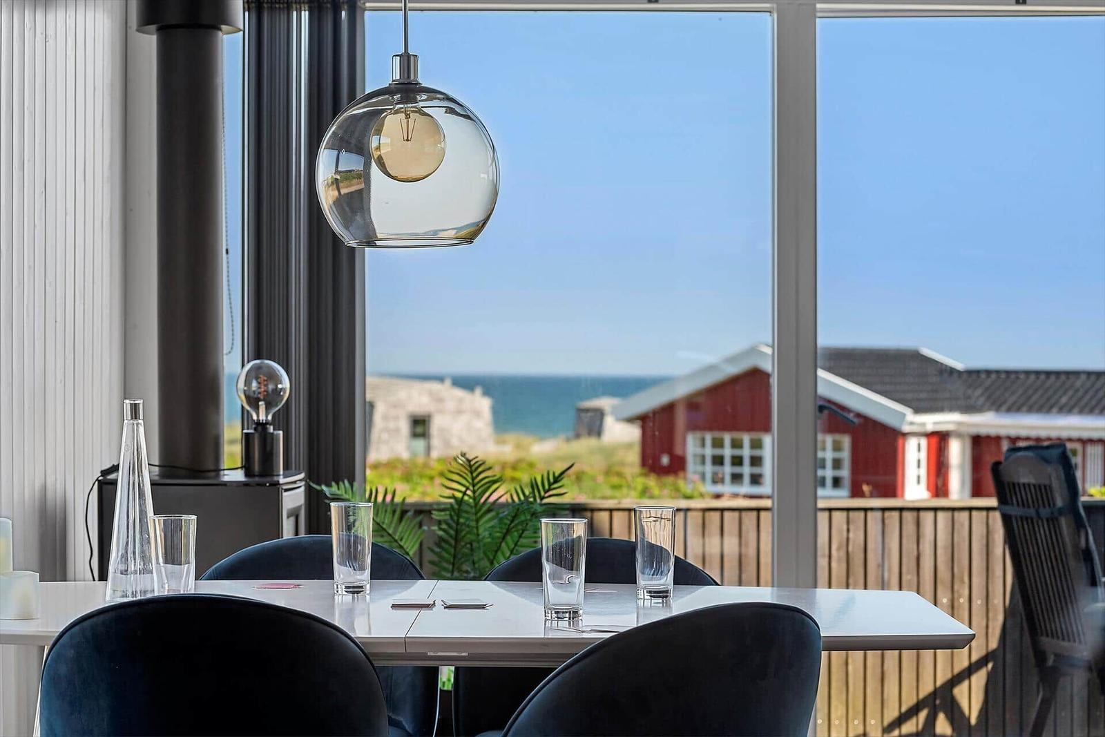 Esstisch mit Stühlen vor großem Fenster mit Meerblick und rotem Haus.