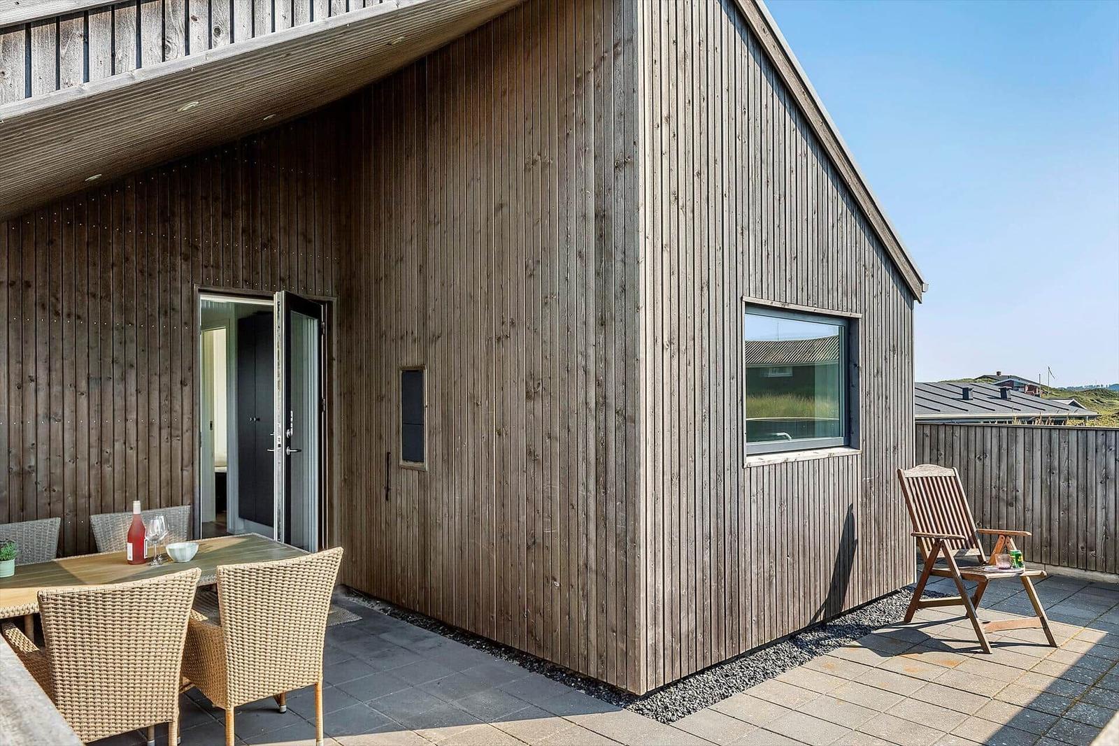 Holzhaus mit Terrasse, Tisch, Stühlen und Sitzgelegenheit. Durchgang zur Wohnung.