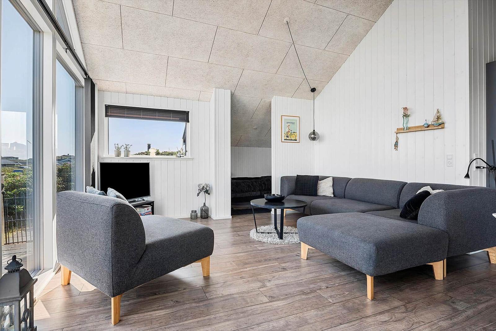 Wohnzimmer mit grauem Sofa, Holzboden und großen Fenstern. An der Wand hängt ein Bild.