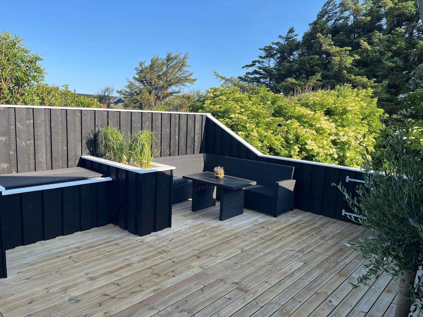 Terrasse mit schwarzer Lounge, Tisch und Pflanzen. Holzdielen und hoher Zaun.