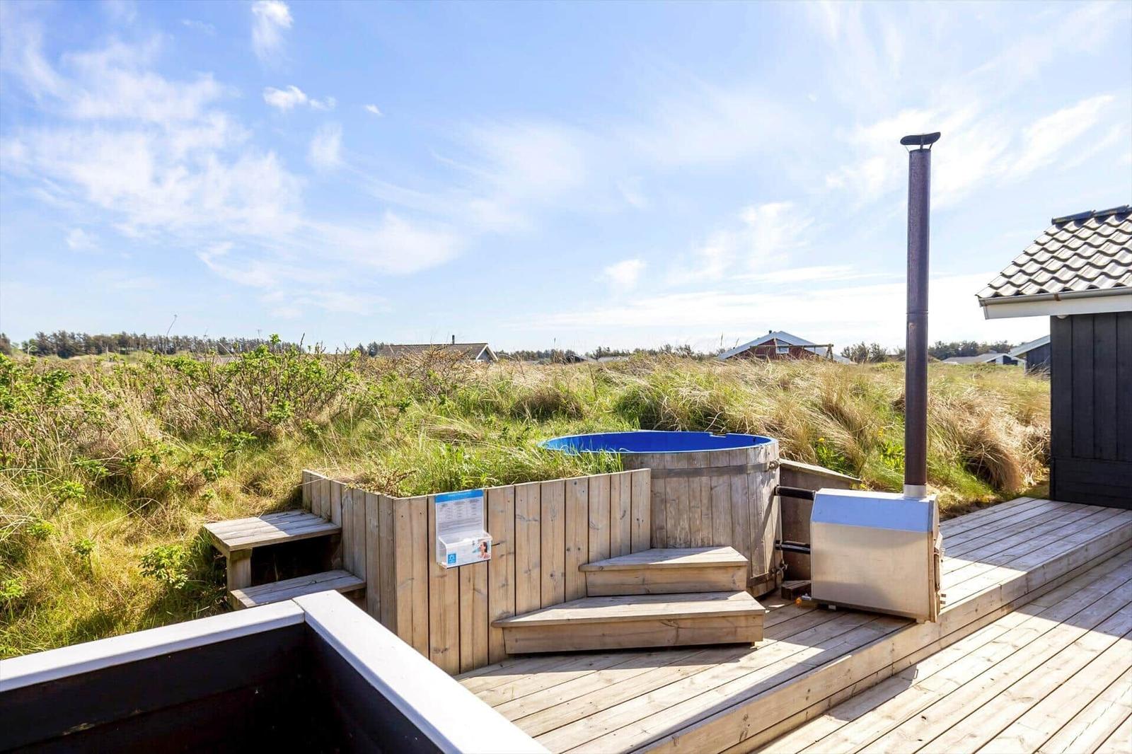 Holzterrasse mit Whirlpool und Grill, umgeben von Gras und Himmel.