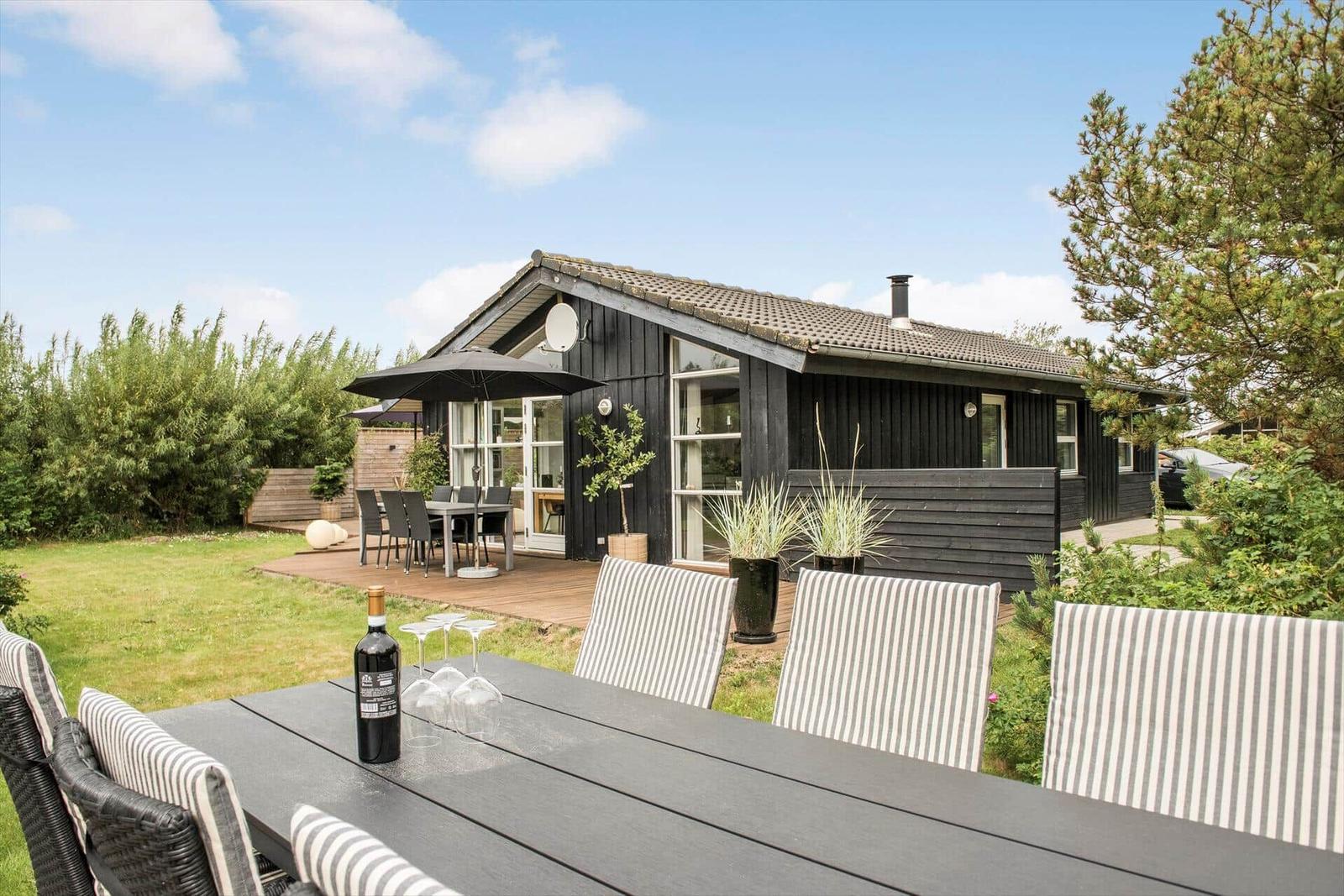 Schwarzes Holzhaus mit Terrasse und Garten. Tisch mit Stühlen und Weinflasche.