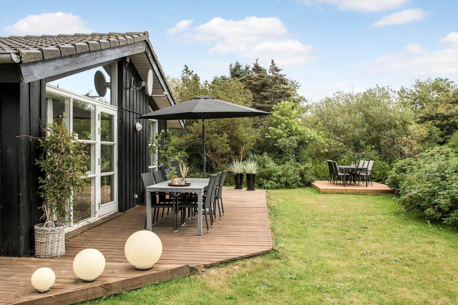 Holzterrasse mit Tisch, Stühlen und Sonnenschirm. Grünflächen und Bäume im Hintergrund.