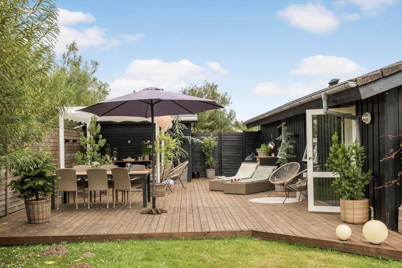 Terrasse mit Tisch, Stühlen, Sonnenliegen und Sonnenschirm. Schwarze Holzwände und Pflanzen.