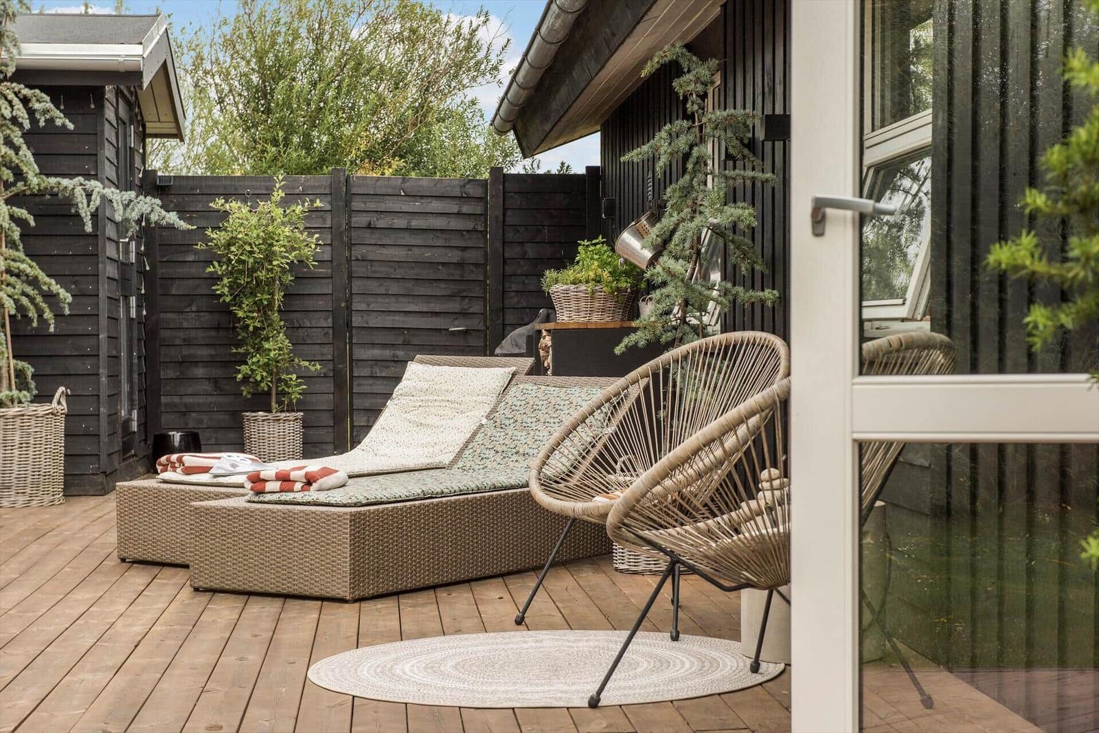 Terrasse mit Liege, Sessel und Pflanzen vor schwarzem Holzhaus.
