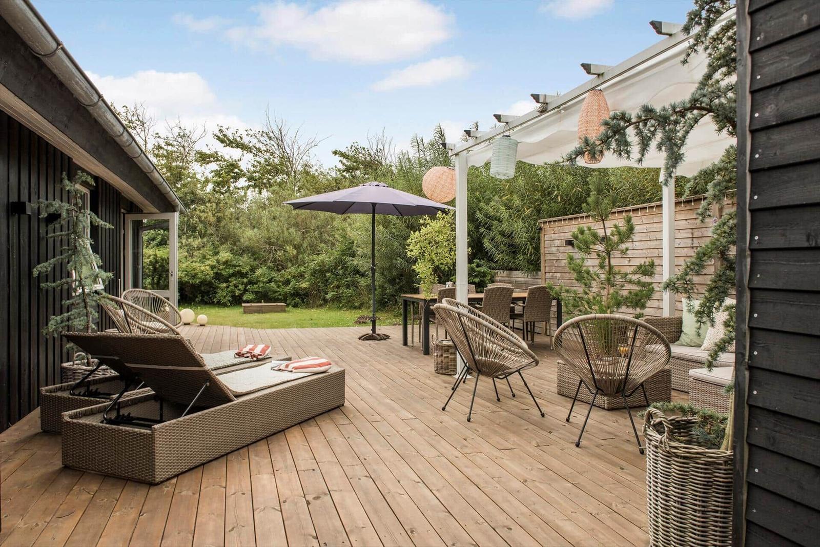 Terrasse mit Liegen, Tisch, Stühlen und Sonnenschirm. Hinterhof mit Grünfläche und Bäumen.