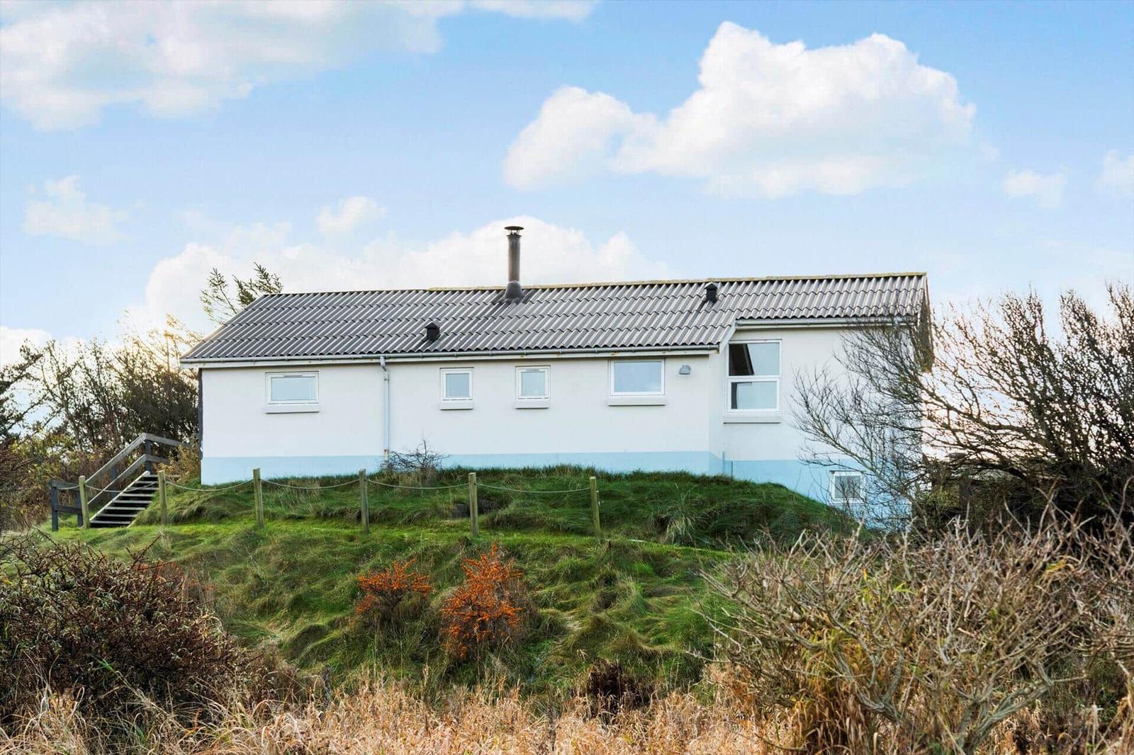 A white house with a gray roof stands on a green hill. A staircase leads to the entrance door.