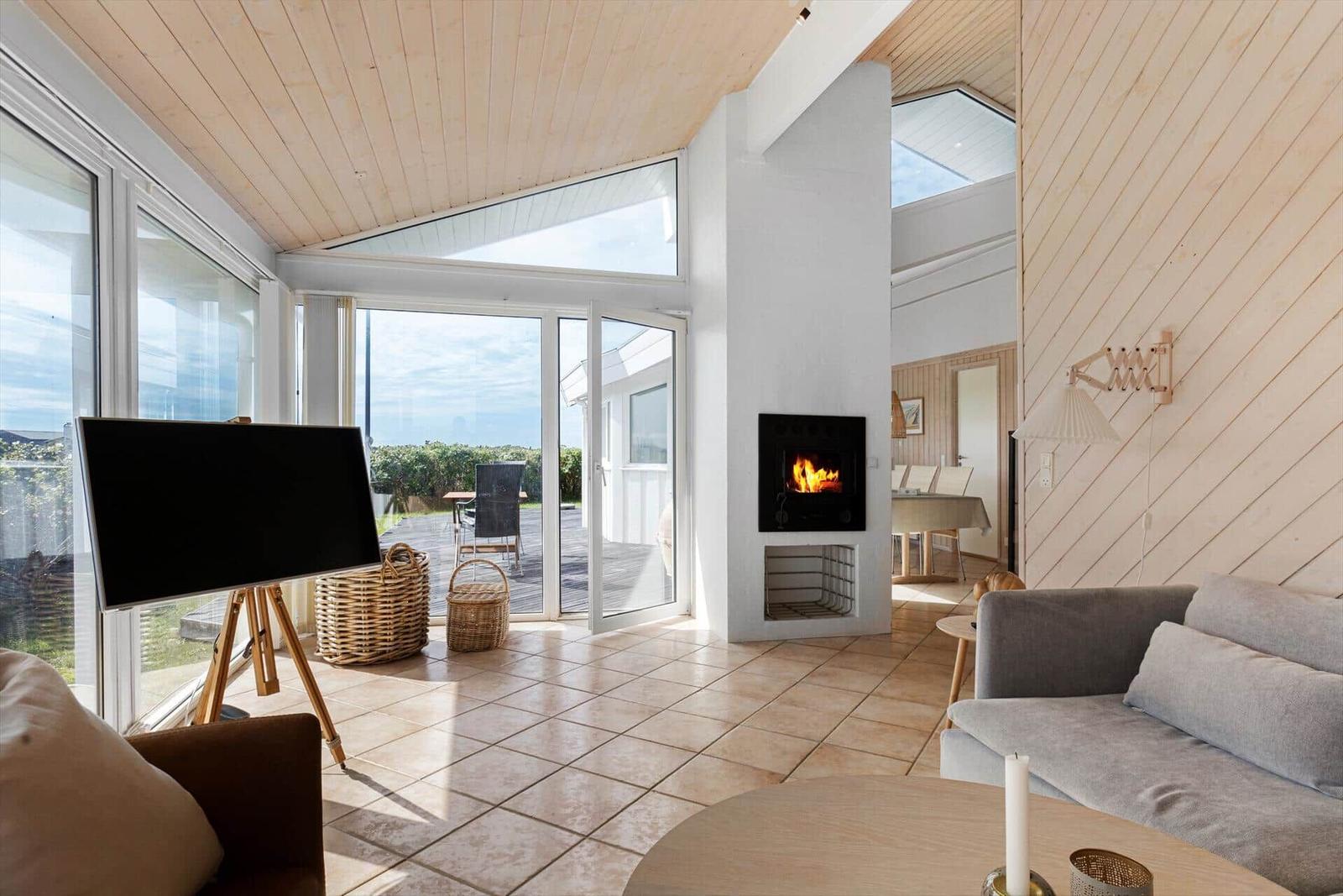 Wohnzimmer mit Kamin, großem Fenster und Terrasse. Holzwände und Fliesenboden.