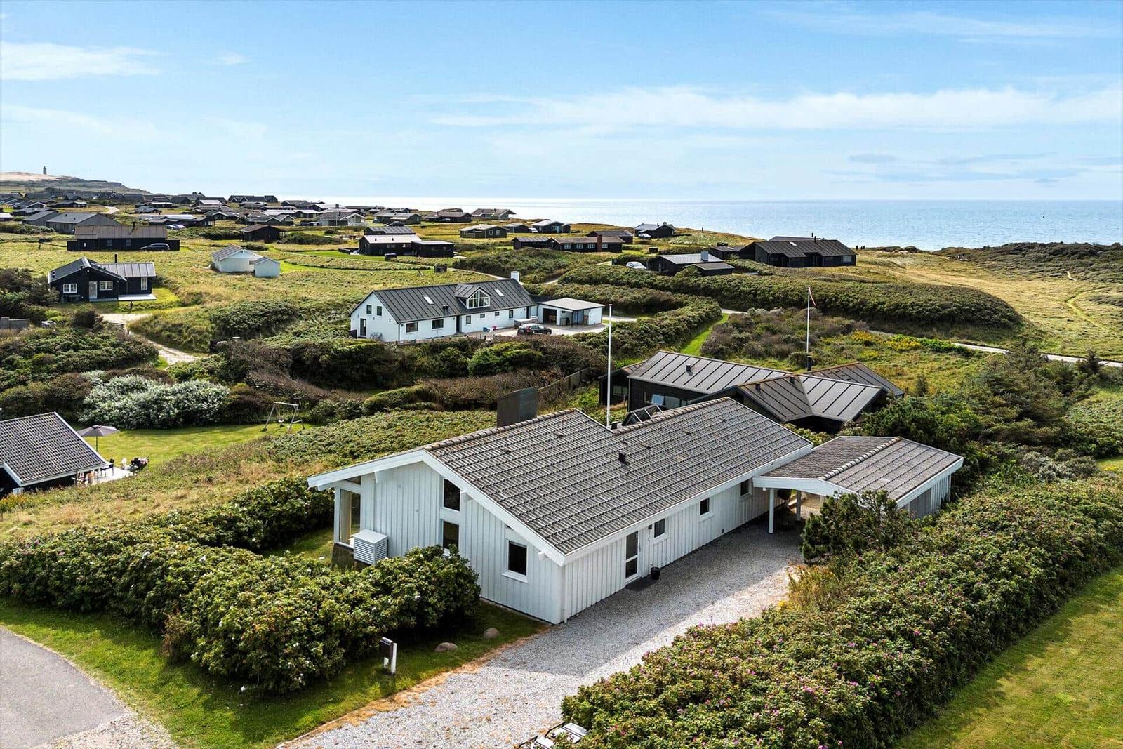 Wohnhaus mit grauem Dach und weißen Wänden, umgeben von Grünflächen und weiteren Häusern nahe dem Meer.