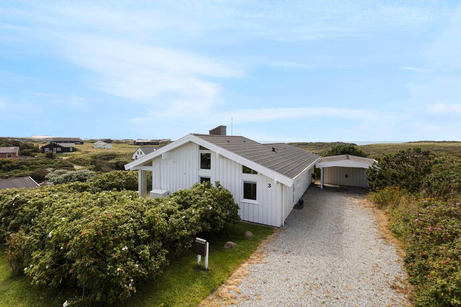 Weißes Ferienhaus mit Garage, umgeben von Grünflächen und Sträuchern unter blauem Himmel.