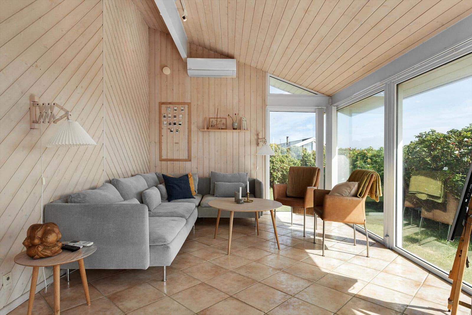 Wohnraum mit Holzwand, Sofa, Tischen und Sesseln. Große Fenster mit Blick auf Garten.