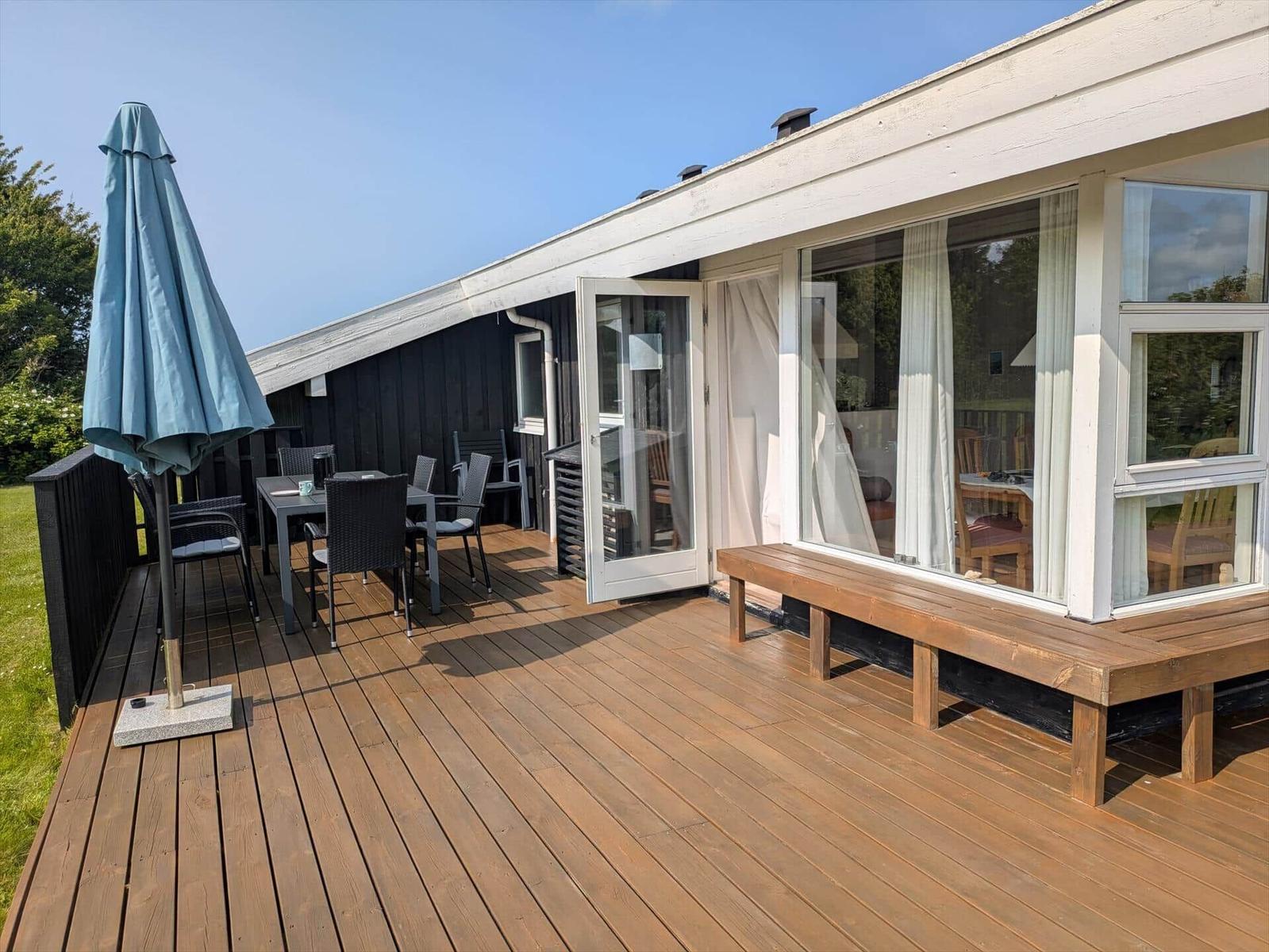 Terrasse mit Tisch, Stühlen und geschlossenem Sonnenschirm. Blick ins Innere durch Fenster.