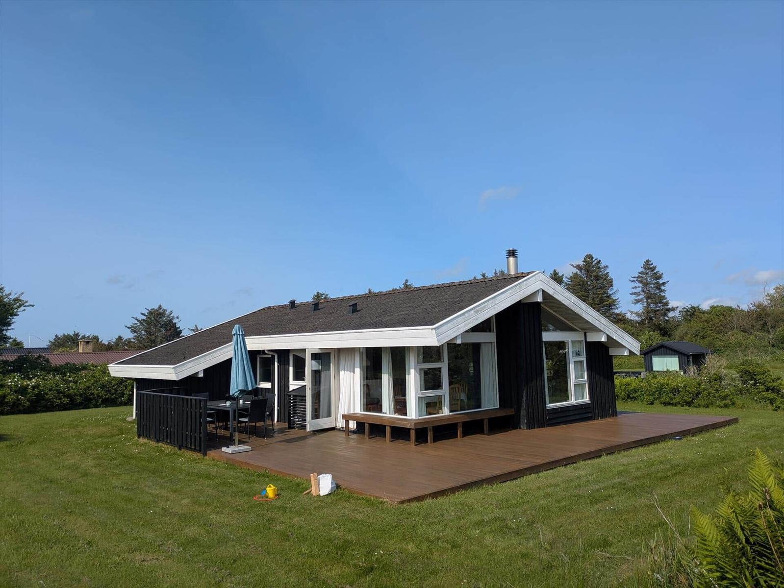 Moderne Holzhütte mit Terrasse, Garten und Gartenmöbeln unter blauem Himmel.