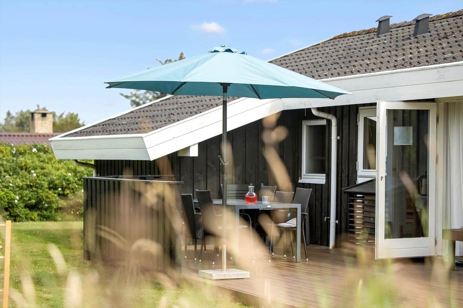 Terrasse mit Tisch, Stühlen und Sonnenschirm. Hintergrund: Haus mit offener Tür und Rasen.