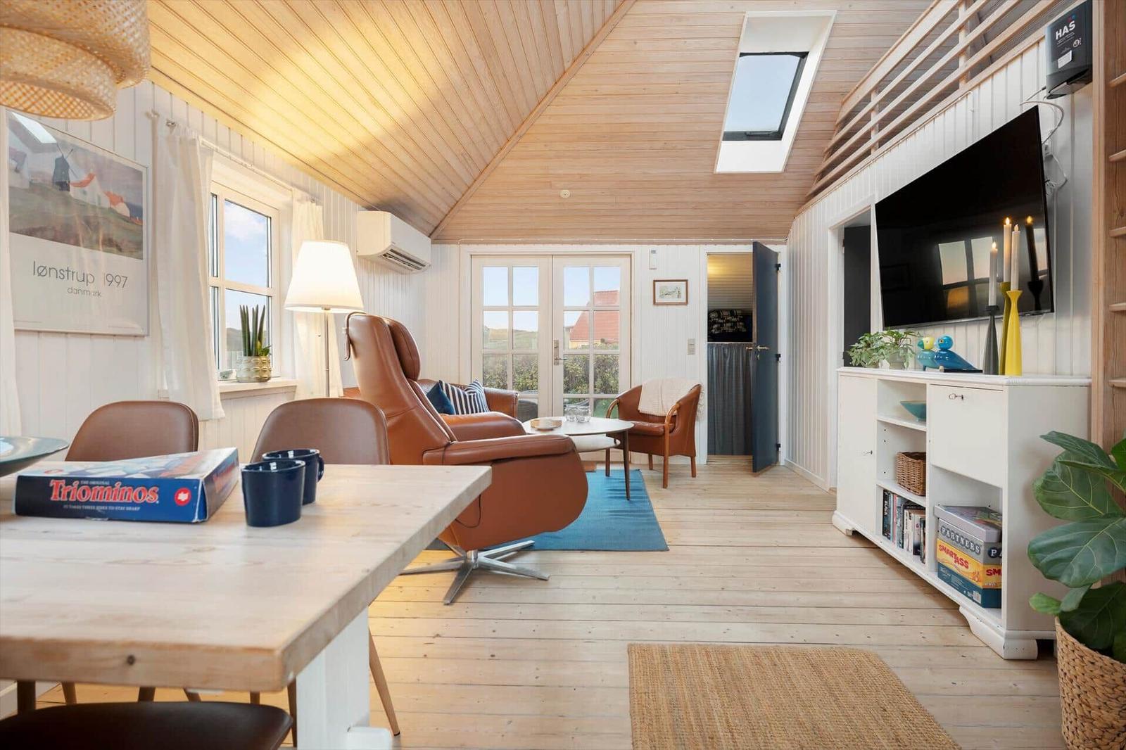 Wohnraum mit Holzdecke, Sofa, Fernseher und Tisch. Fenster und Tür zur Außenseite.