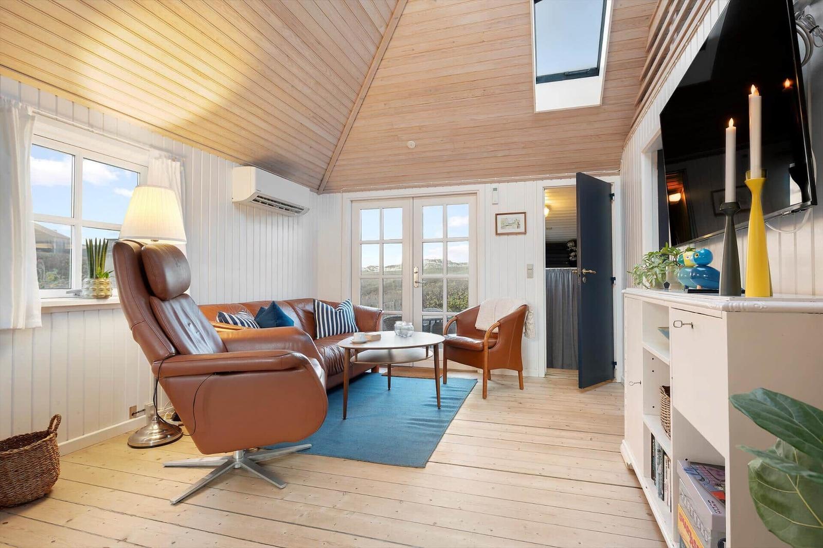 Wohnraum mit braunen Ledermöbeln, Holzdecke und Fenstern. TV, Bücherregal und Kamin.